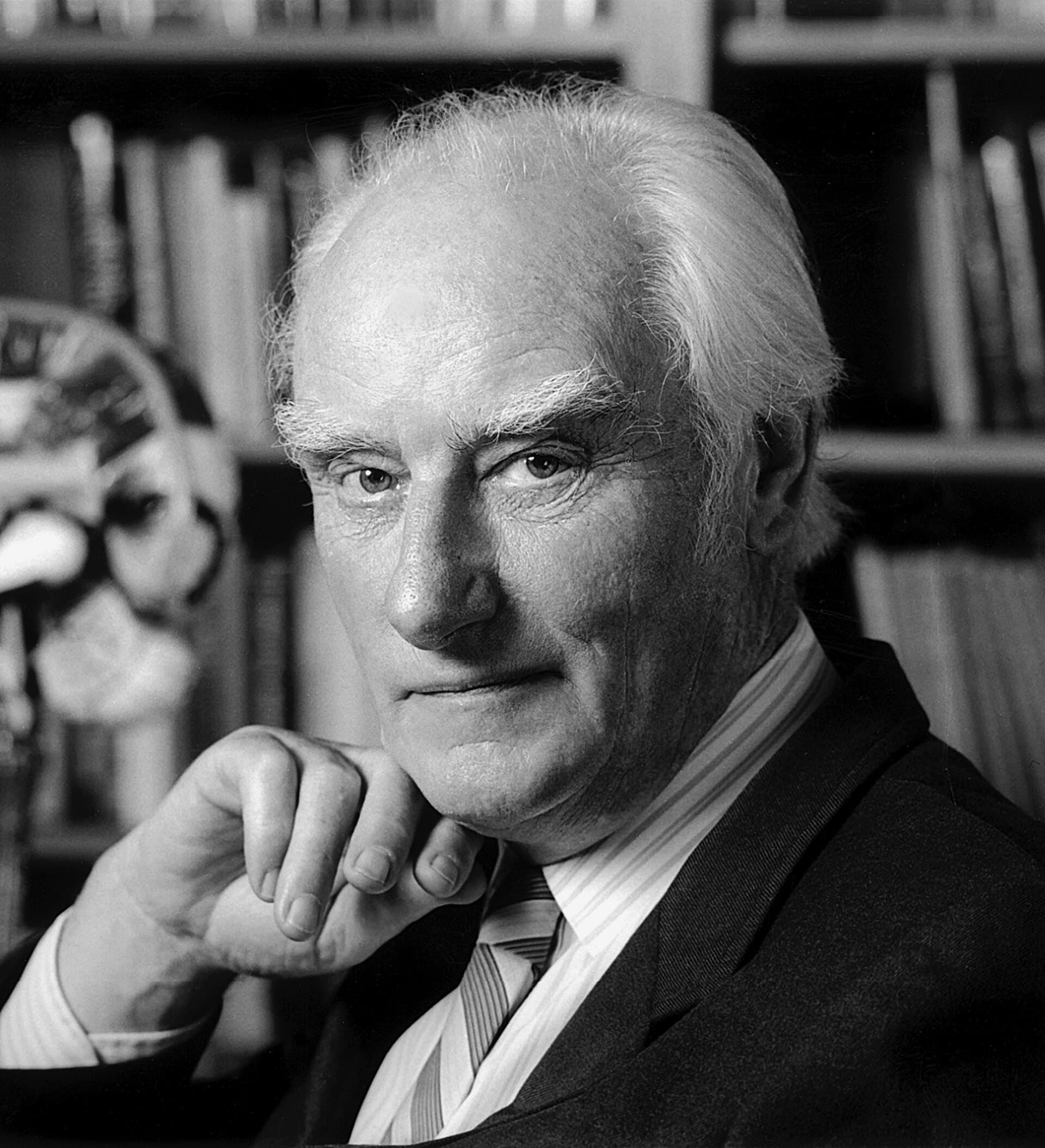 Francis Crick, white-haired, resting his chin on his hand in front of bookshelves.