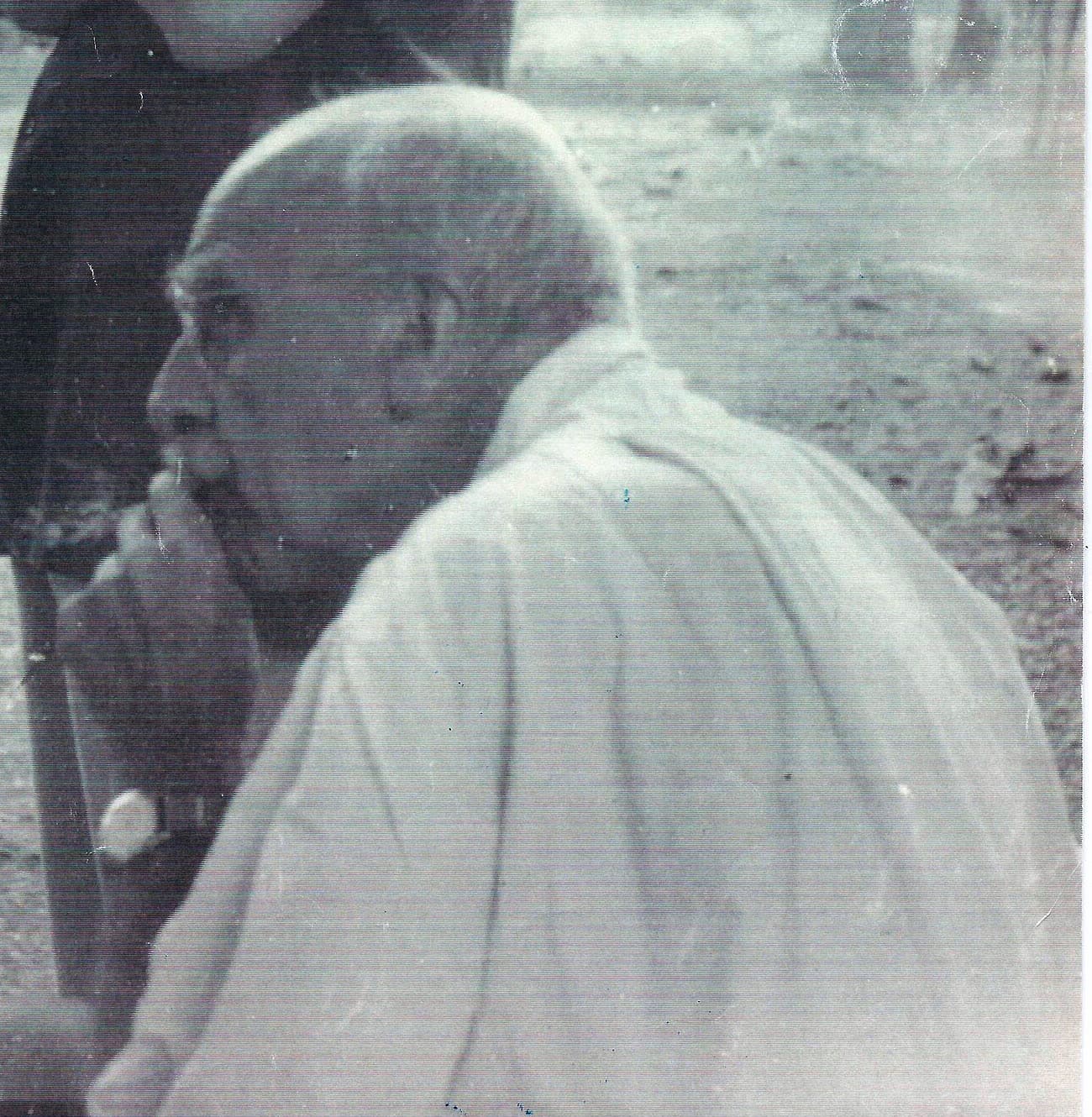 J. B. S. Haldane, an old man in a white shirt, seated outdoors with his hand at his chin.