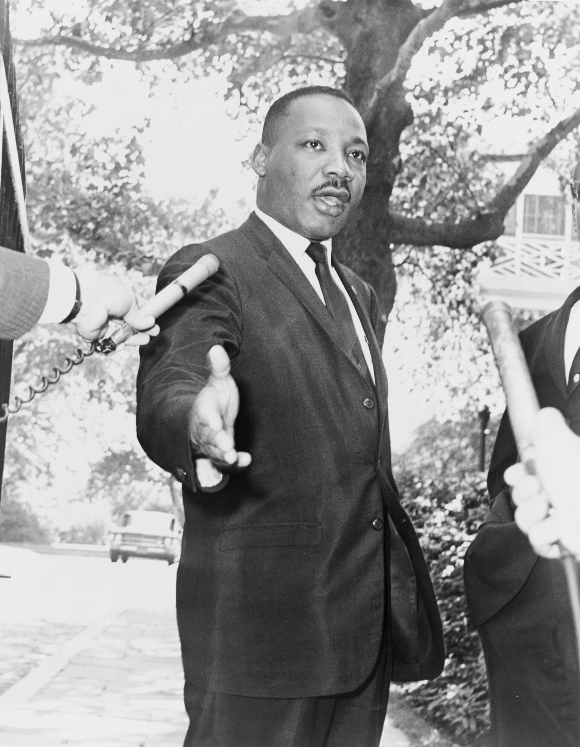 Martin Luther King Jr. in a dark suit, gesturing with one hand at a press conference.