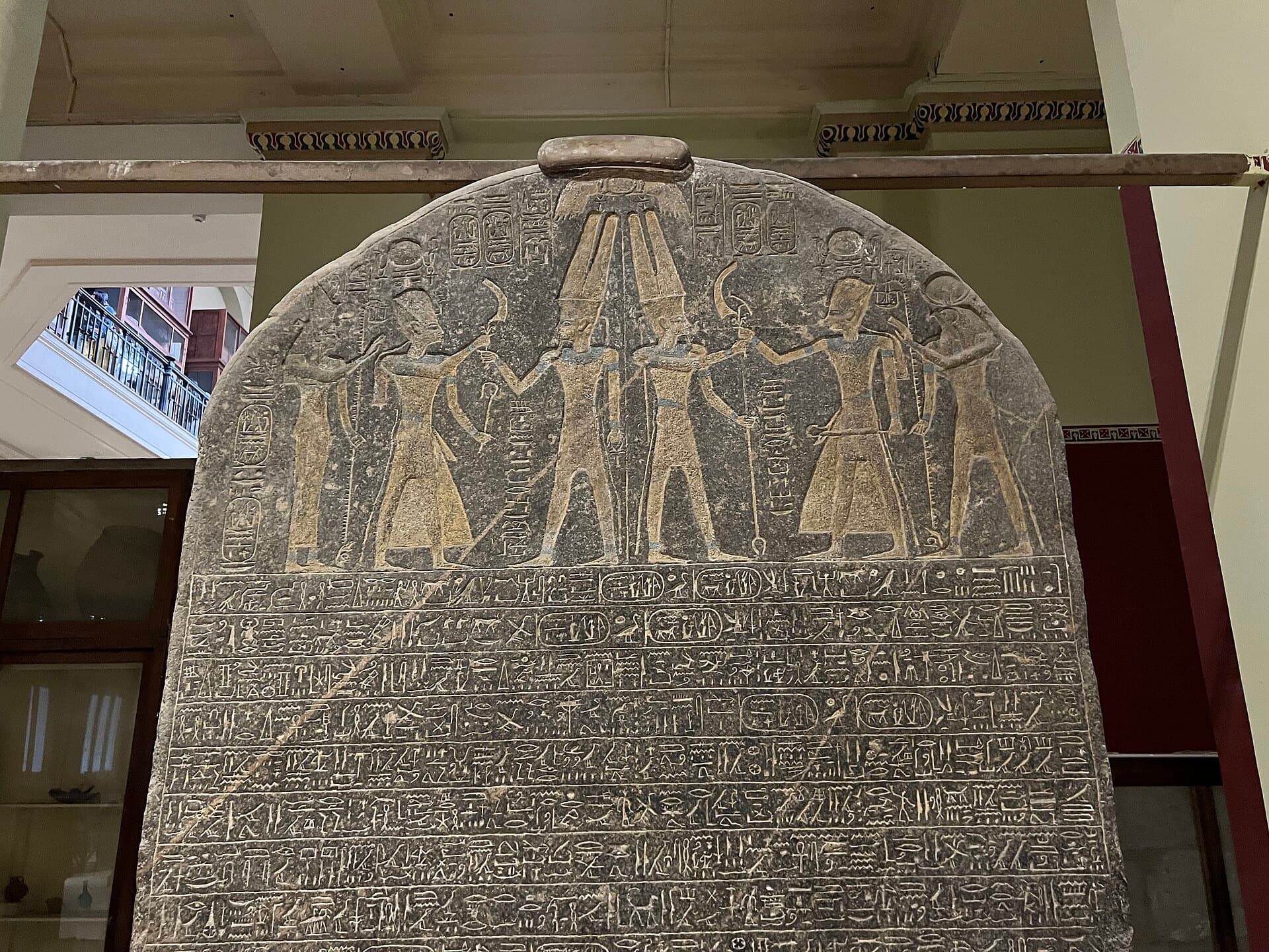 A tall rounded black granite slab carved with figures of a pharaoh and gods above rows of hieroglyphs.
