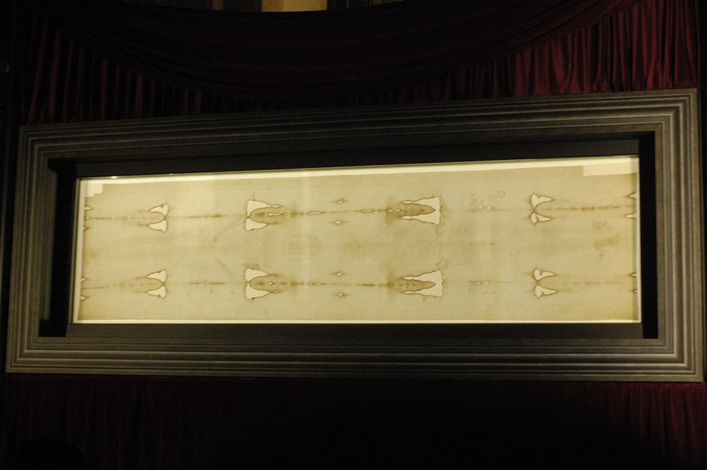 A long, pale linen cloth in a dark frame behind glass, marked with faint brown stains and rows of pale patches.